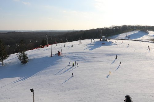 A snowy ski slope with skiers and snowboarders, surrounded by trees and a ski lift under a clear sky.