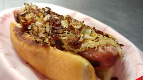 A grilled hot dog topped with sautéed onions and sauerkraut in a soft bun, served on a pink paper liner.