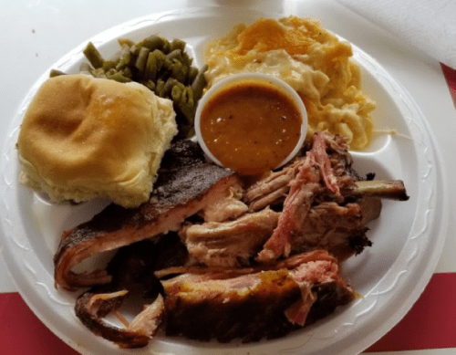 A plate of barbecue ribs, green beans, macaroni and cheese, a roll, and a small cup of sauce.