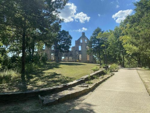 Ruins of a stone building surrounded by trees, with a clear blue sky and a pathway leading through the landscape.