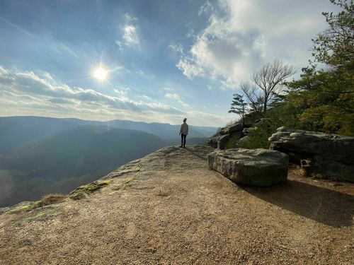 A person stands on a rocky ledge, overlooking a valley with mountains under a bright sky and soft clouds.
