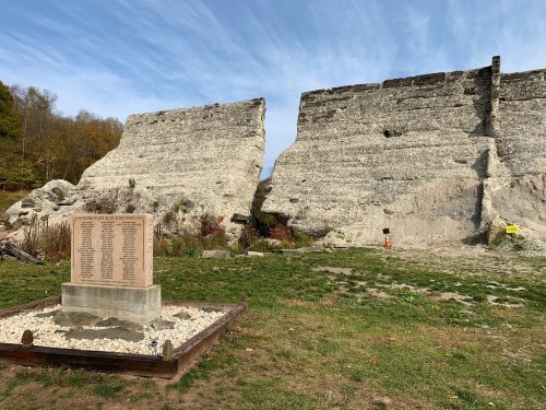 A weathered stone wall with a memorial plaque in front, surrounded by grass and trees under a blue sky.
