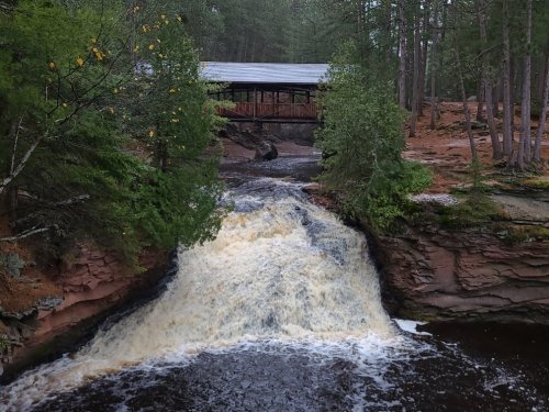A waterfall cascades over rocky terrain, with a wooden bridge and lush greenery in the background.