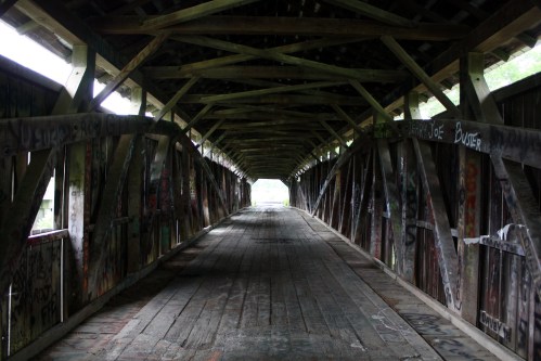 A dimly lit covered bridge interior with wooden beams and graffiti on the walls, leading to a bright exit.
