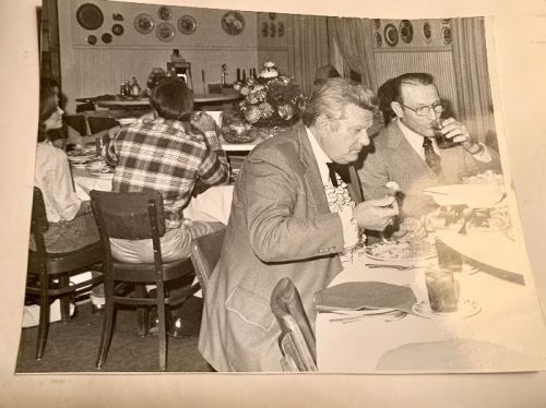 Black and white photo of men dining at a restaurant, with plates of food and drinks on the table.