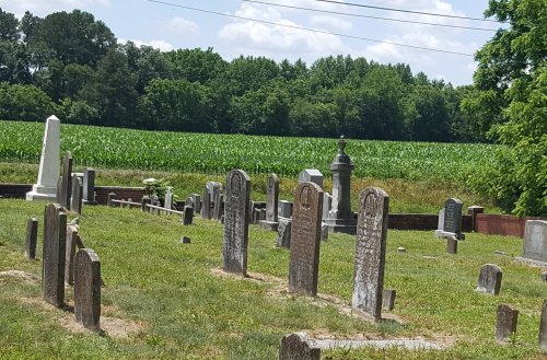A cemetery with various gravestones, surrounded by green fields and trees under a blue sky with clouds.