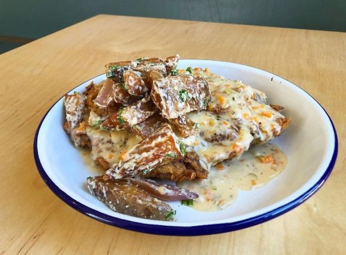 A plate of crispy fried chicken topped with creamy gravy and garnished with herbs, served with fried potato wedges.
