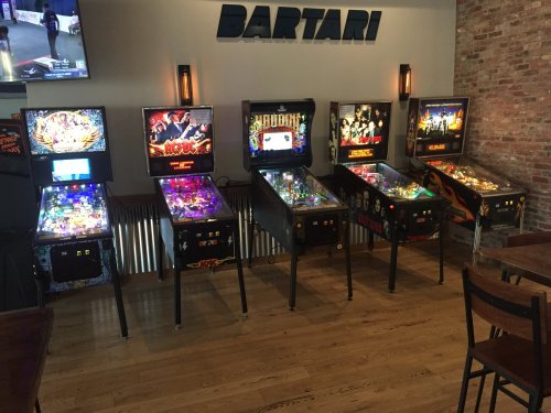 A row of colorful pinball machines in a bar setting, with a brick wall and a TV in the background.