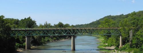 A green metal bridge spans a river, surrounded by lush trees and hills under a clear blue sky.