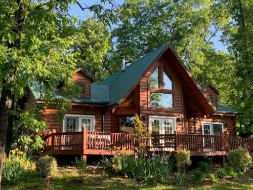 A charming log cabin surrounded by lush greenery and colorful flowers, featuring a large deck and a sloped roof.