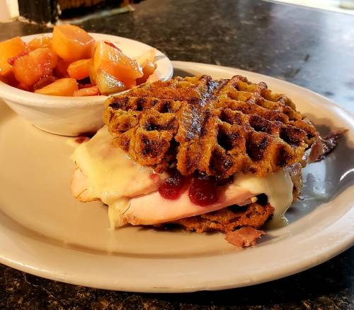 A savory sandwich made with a waffle, turkey, cheese, and cranberry sauce, served with a side of fruit.