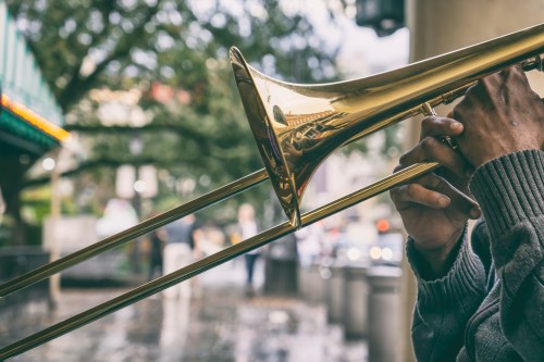 A close-up of a person playing a trombone, with a blurred city street in the background.
