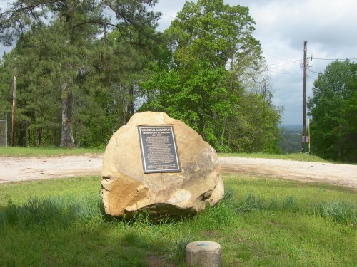 A large rock with a plaque stands in a grassy area, surrounded by trees and a dirt road in the background.
