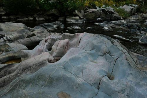 Smooth, multicolored rock formations beside a calm river, surrounded by greenery and scattered boulders.