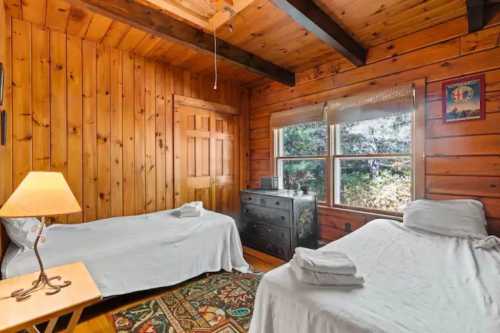 Cozy wooden bedroom with two twin beds, a dresser, and a window overlooking greenery. Warm, rustic decor.