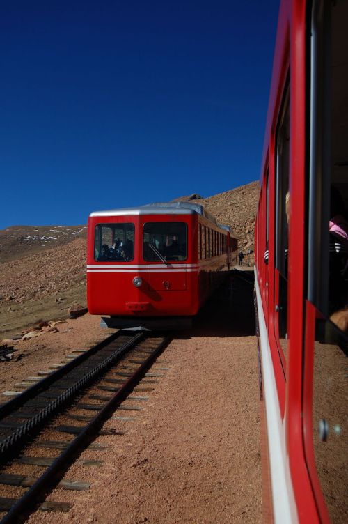 A red train approaches on a track beside a rocky landscape under a clear blue sky.