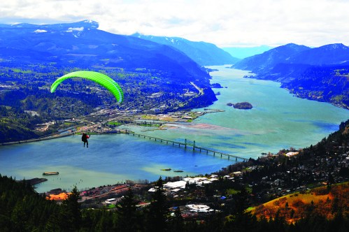 A paraglider soaring above a river valley, surrounded by mountains and lush greenery, with a bridge in the distance.