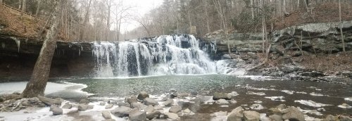 A serene waterfall cascades over rocks into a frozen pool, surrounded by bare trees and winter scenery.