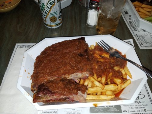 A plate of ribs topped with chili, served with a side of fries and a drink in the background.