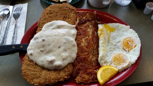 A hearty breakfast plate featuring fried chicken, creamy gravy, hash browns, and two sunny-side-up eggs with lemon slices.