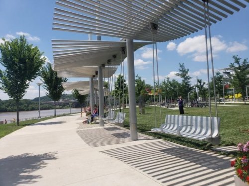 A park with modern swings under a metal canopy, surrounded by trees and a river in the background on a sunny day.