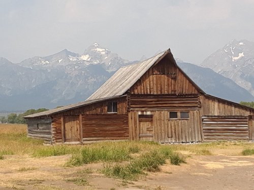 A rustic wooden barn stands in a grassy field with snow-capped mountains in the background under a clear sky.