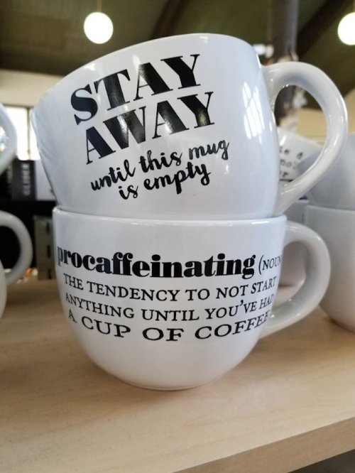 Two stacked coffee mugs with playful text: "STAY AWAY until this mug is empty" and "procaffeinating: the tendency to not start anything until you've had a cup of coffee."