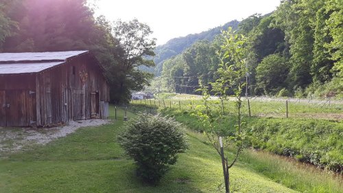 A rustic wooden barn beside a grassy area, with trees and a winding road in a lush green landscape.