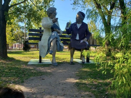 Two life-sized statues of women sitting on a park bench, engaged in conversation amidst trees and greenery.