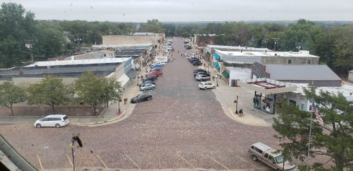 Aerial view of a small town street lined with shops and parked cars, featuring cobblestone pavement and trees.