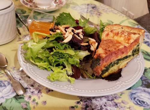 A plate with a slice of quiche, mixed greens salad, and a small cup of orange jelly on a floral tablecloth.