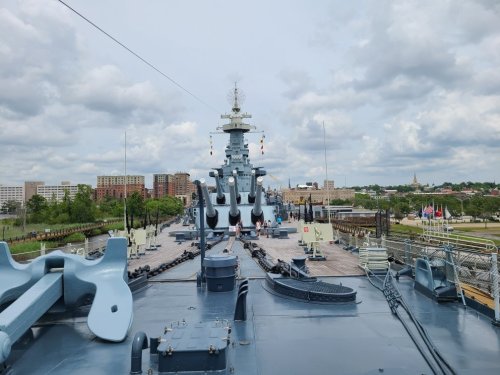 View from the deck of a battleship, showcasing the ship's bow and city skyline under a cloudy sky.