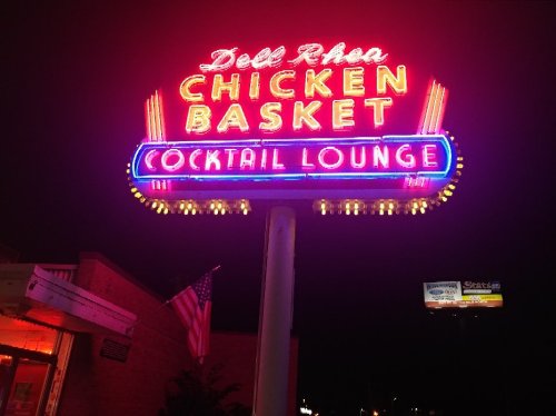 Neon sign for Dell Rhea Chicken Basket Cocktail Lounge, glowing brightly at night with an American flag nearby.