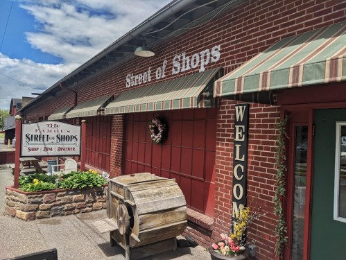 Exterior of a brick building with a sign reading "Street of Shops," featuring a welcome sign and flower decorations.