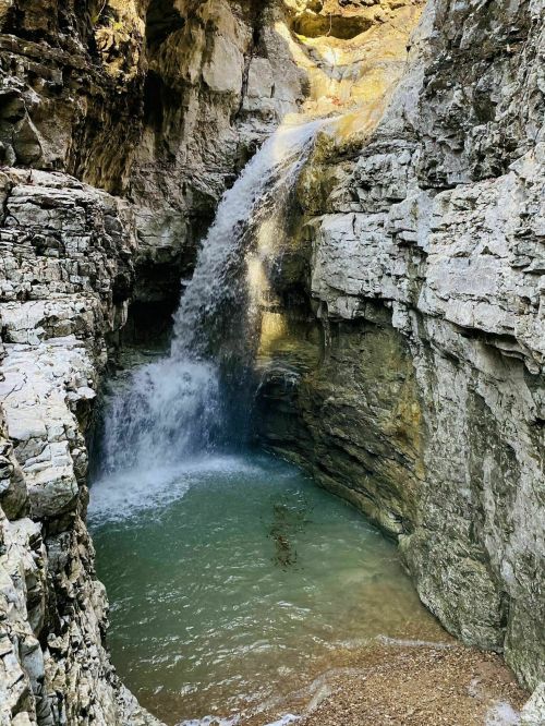 A serene waterfall cascades into a turquoise pool, surrounded by rugged stone cliffs in a natural setting.