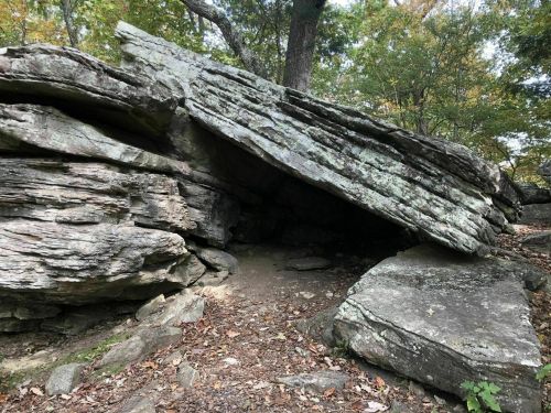 A large, flat rock formation partially overhangs a dirt path, surrounded by trees and foliage.