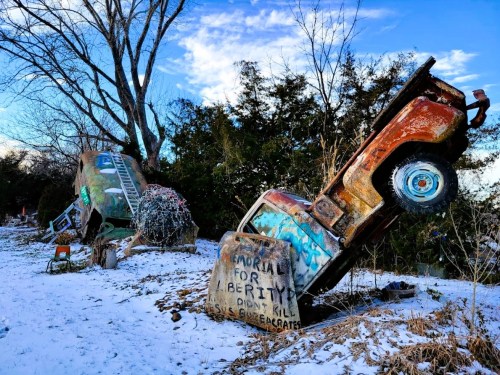 Two rusted vehicles, one upright and one tilted, surrounded by snow and trees, with graffiti on their surfaces.