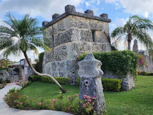 Stone tower surrounded by greenery and palm trees, with a sign reading "ADMIO: DROP BELOW." in a landscaped area.