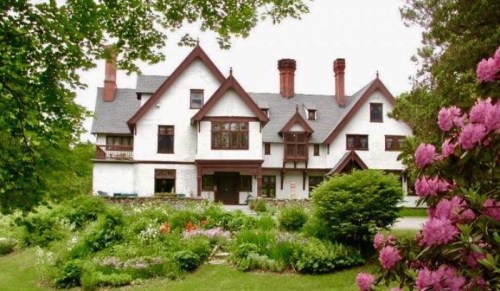 A large, elegant white mansion with red accents, surrounded by lush greenery and colorful flowers.
