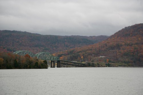 A green bridge spans a river, surrounded by autumn-colored hills under a cloudy sky.