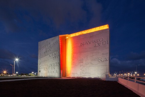 A modern concrete monument illuminated in orange at dusk, featuring engraved text and a sleek design.