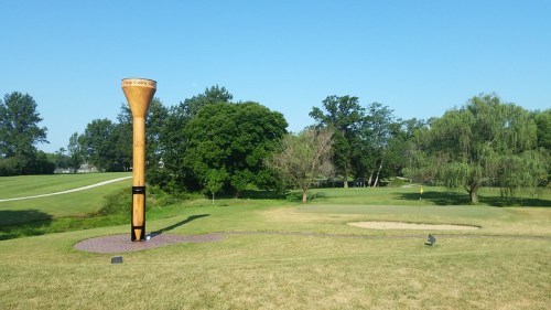 A large wooden golf tee sculpture stands on a grassy golf course near a sand trap and trees under a clear blue sky.