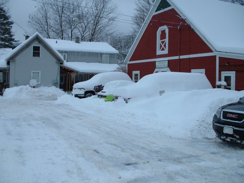 Snow-covered street with parked cars and houses, blanketed in white snow under a gray sky.