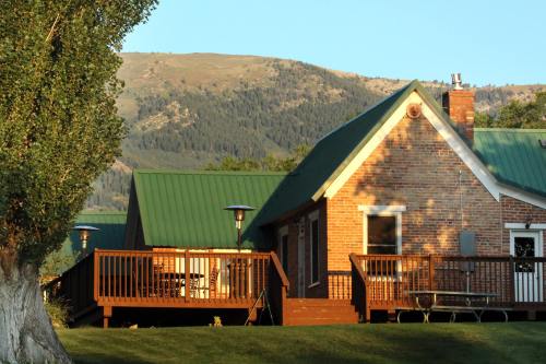 A brick house with a green metal roof, surrounded by trees and mountains in the background, under a clear blue sky.