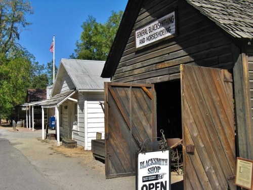 A rustic blacksmith shop with open doors, surrounded by wooden buildings and trees, under a clear blue sky.
