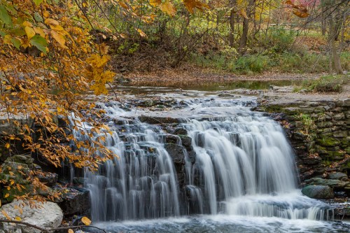 A serene waterfall cascades over rocks, surrounded by autumn foliage in vibrant shades of orange and yellow.