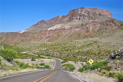 A winding road leads through a desert landscape with rocky mountains under a clear blue sky.