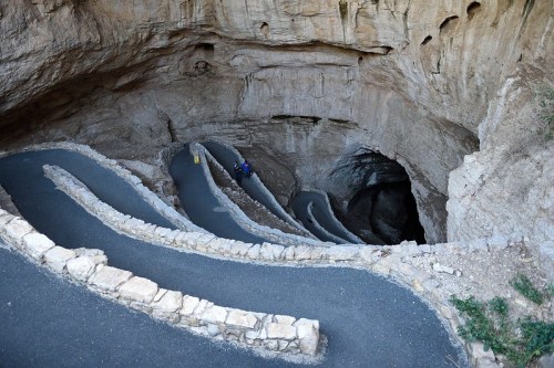 Winding path inside a cave, leading down into a dark opening, with rocky walls and a few people visible.