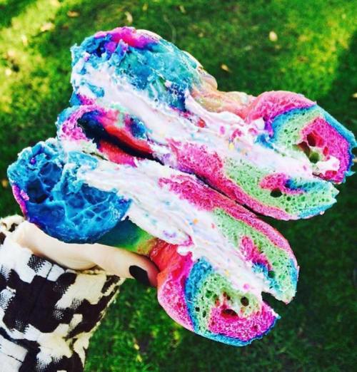 A hand holding a colorful, rainbow bagel filled with cream cheese and sprinkles, set against a grassy background.