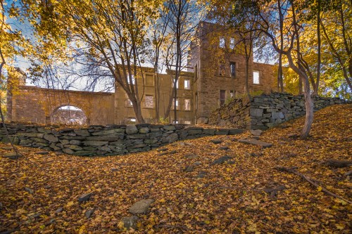 Ruins of a stone building surrounded by autumn foliage and trees, with a carpet of fallen leaves on the ground.